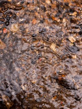 the rippled surface of running water with multicolored natural pebbles below the water surface and some sticking out shining and glistening as water travels over and down stream in the day light