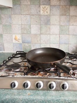 A Big Black Round Circular Frying Pan without Any Food on the Top of Gas Cooker hob with Dials all Turned off, and with bits of dirt and burnt food on some foil underneath the metal frame and with tiles in the background in uk kitchen