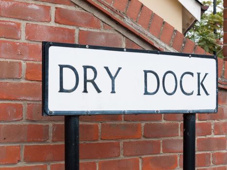 a black and white street sign saying dry dock with brick background attached to poles into the ground on the path and road leading the way naming uk england sailing community seafront