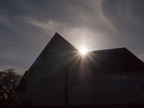 the silhouette of a pointy house with a sun flare peaking through in the middle top part through the sky sun setting cool effect in spring and summer