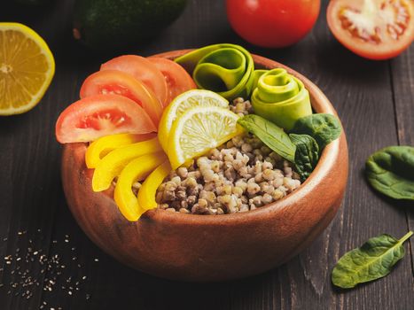 Close up view of vegetarian buddha bowl with green buckwheat, spinach, avocado, tomatoes and yellow sweet pepper paprika on dark wooden table with copy space
