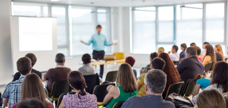 Business and entrepreneurship symposium. Speaker giving a talk at business meeting. Audience in conference hall. Rear view of unrecognized participant in audience.