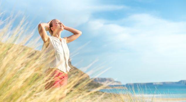 Relaxed woman, arms rised, enjoying sun, freedom and life an a beautiful beach. Young lady feeling free, relaxed and happy. Concept of vacations, freedom, happiness, enjoyment and well being.