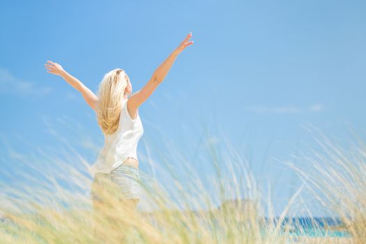 Relaxed woman, arms raised, enjoying sun, freedom and life an a beautiful beach. Young lady feeling free, relaxed and happy. Concept of vacations, freedom, happiness, enjoyment and well being.