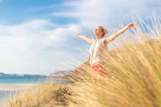 Relaxed woman, arms rised, enjoying sun, freedom and life an a beautiful beach. Young lady feeling free, relaxed and happy. Concept of vacations, freedom, happiness, enjoyment and well being.