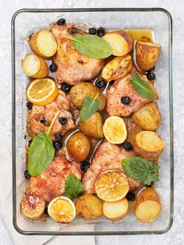 Top view of chicken thighs with potatoes, lemon and black olives, cooked in oven on gray concrete background. Baked chicken leg quarter in heat-proof glass. Vertical.