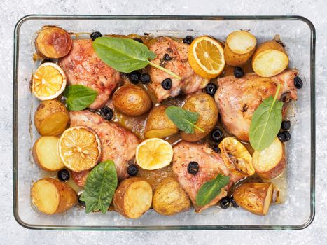 Top view of chicken thighs with potatoes, lemon and black olives, cooked in oven on gray concrete background. Baked chicken leg quarter in heat-proof glass.