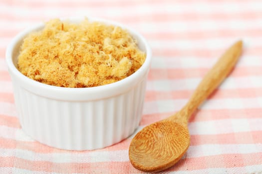 dried pork floss in white cup with wooden spoon on napery background.  
