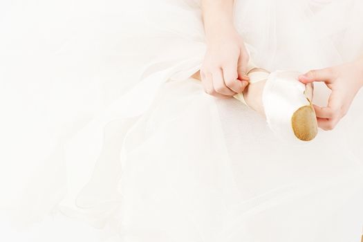 A small ballerina dresses or takes off her pointe isolated on white background. Slender little ballerina girl in white dress and in pointe shoes