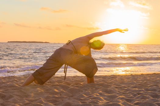 Silhouette of young woman practicing yoga on sandy beach at sunset.