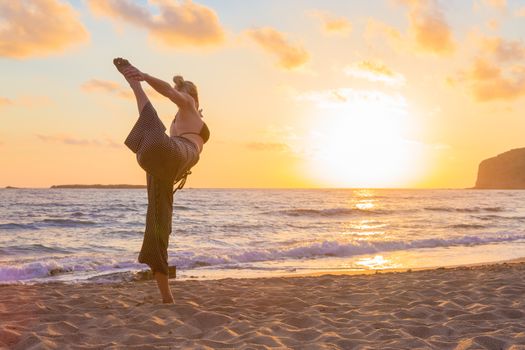 Silhouette of young woman practicing standing bow yoga pose on sandy beach at sunset.