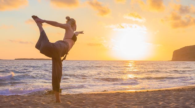 Silhouette of young woman practicing standing bow yoga pose on sandy beach at sunset.