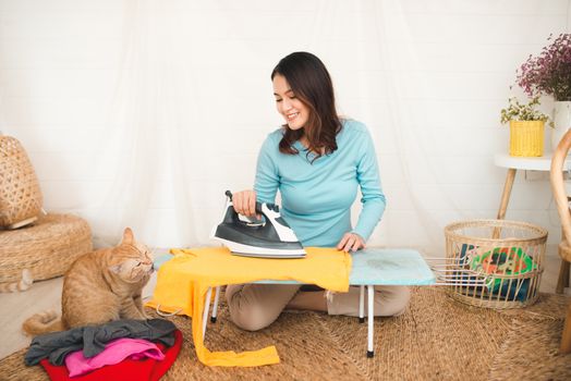 Happy young asian woman ironing clothes sitting on floor at home.