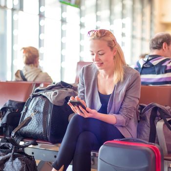Casual blond young woman using her cell phone while waiting to board a plane at the departure gates. Wireless network hotspot enabling people to access internet conection. Public transport.