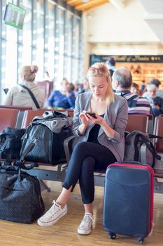 Casual blond young woman using her cell phone while waiting to board a plane at the departure gates. Wireless network hotspot enabling people to access internet conection. Public transport.