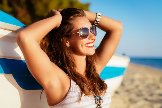 Beautiful young woman relaxing on the beach. She is sitting next to boat and enjoying at sunbathing.