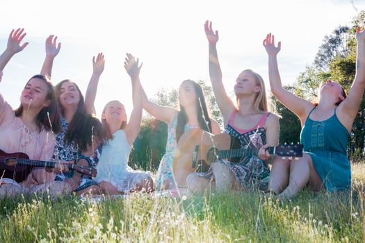 Young Girls Sitting Together in Green Grassy Field Singing and Playing Musical Instruments With Arms Raised up