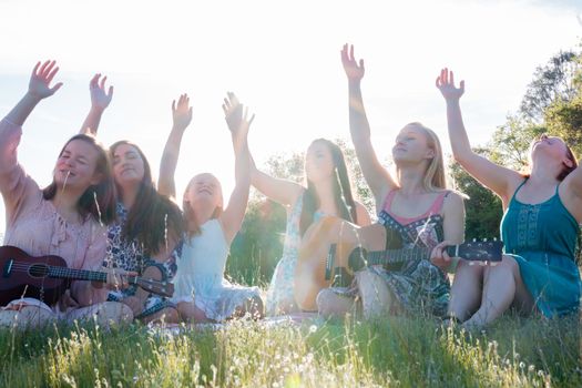 Young Girls Sitting Together in Green Grassy Field Singing and Playing Musical Instruments With Arms Raised up