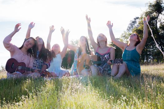 Young Girls Sitting Together in Green Grassy Field Singing and Playing Musical Instruments With Arms Raised up