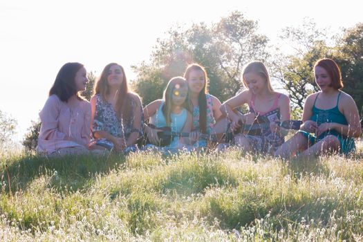 Young Girls Sitting Together in Grassy Field Singing and Playing Musical Instruments With Sunlight Overhead