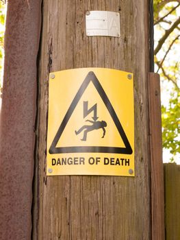 a yellow and black danger of death sign with triangle and lightening bolt on a wooden electric pole pylon outside resource