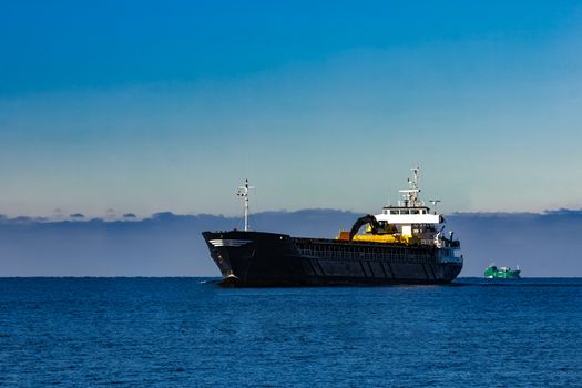 Black cargo ship with long reach excavator moving by baltic sea