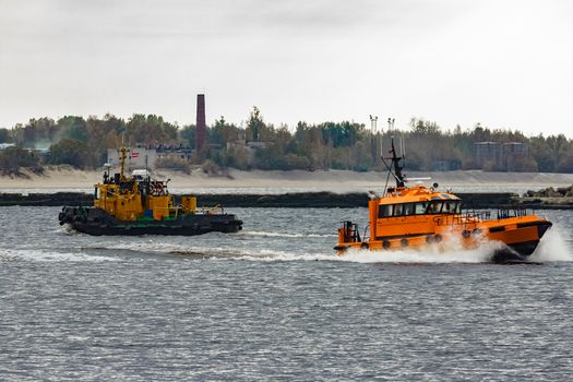 Orange pilot ship moving at speed past the tug ship in Riga