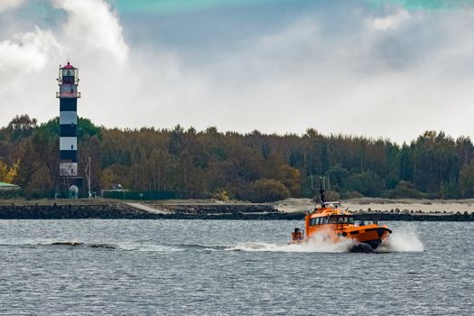 Orange pilot ship moving at speed past the lighthouse in Riga