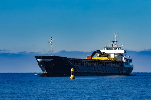 Black cargo ship with long reach excavator moving by baltic sea