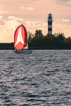 Red sailboat sailing past the big lighthouse in evening, Latvia