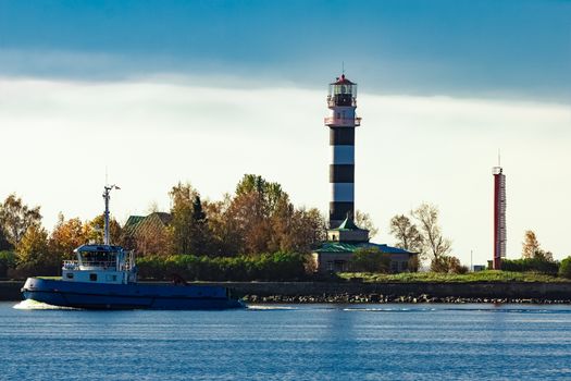 Blue small tug ship sailing past the big lighthouse at sunny day