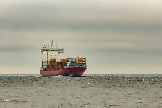 Red cargo container ship sailing from Baltic sea in cloudy day