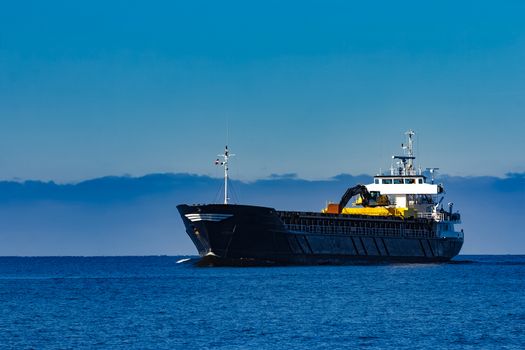 Black cargo ship with long reach excavator moving by baltic sea