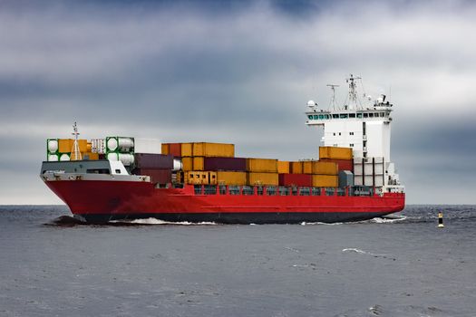 Red cargo container ship sailing from Baltic sea in cloudy day