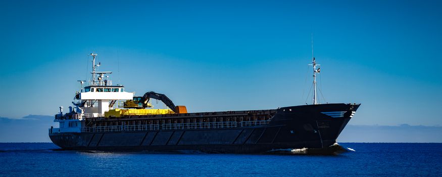 Black cargo ship with long reach excavator moving by baltic sea