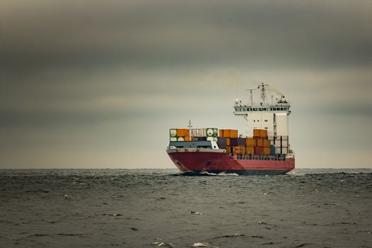 Red cargo container ship sailing from Baltic sea in cloudy day