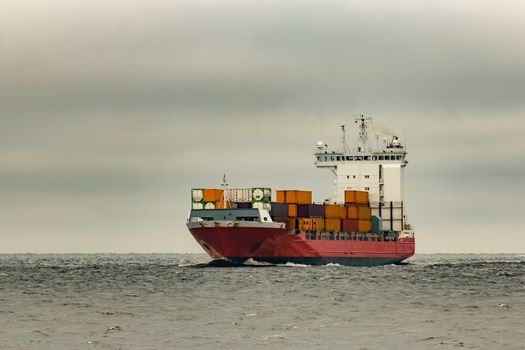 Red cargo container ship sailing from Baltic sea in cloudy day