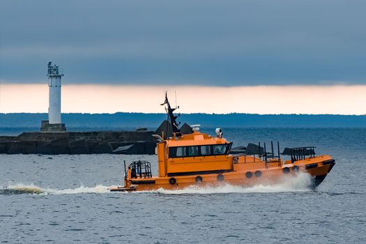 Orange pilot ship moving at speed past the lighthouse in Riga