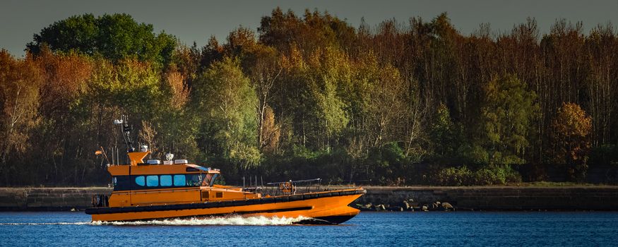 Orange pilot ship sailing past the autumn trees in Europe