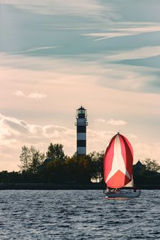 Red sailboat sailing past the big lighthouse in evening, Latvia