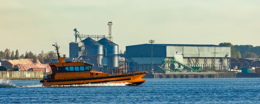 Orange pilot ship sailing past the factory in Latvia