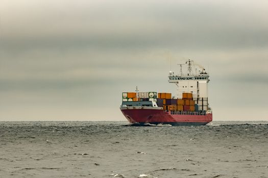 Red cargo container ship sailing from Baltic sea in cloudy day