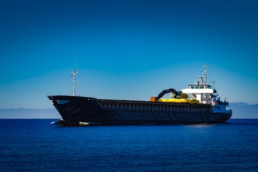 Black cargo ship with long reach excavator moving by baltic sea