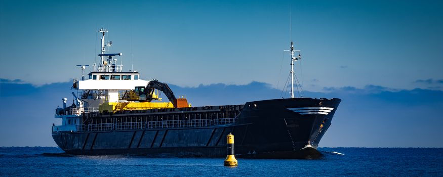 Black cargo ship with long reach excavator moving by baltic sea