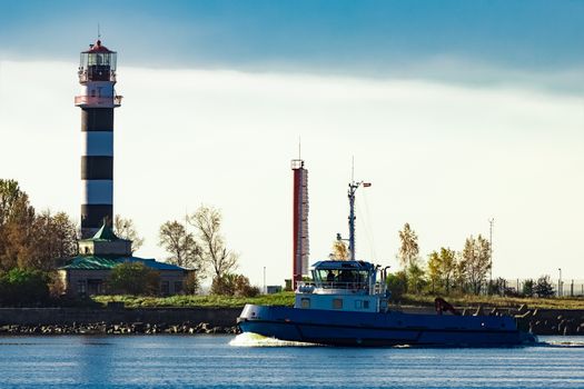 Blue small tug ship sailing past the big lighthouse at sunny day