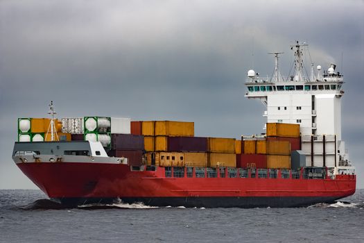 Red cargo container ship sailing from Baltic sea in cloudy day
