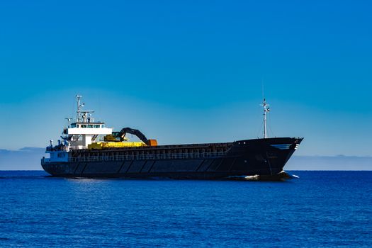 Black cargo ship with long reach excavator moving by baltic sea