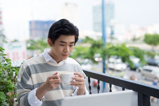 Handsome asian young man working on laptop and smiling while enjoying coffee in cafe