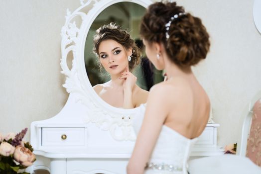 Beautiful young bride in front of the mirror. Portrait with wedding makeup, hairstyle, dress