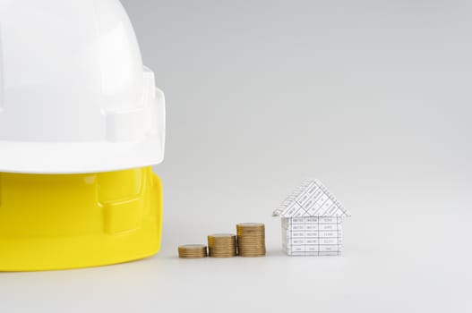 Step pile of gold coins between house and white on yellow engineer hat on white background.